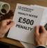 A close-up of a businessperson's hands holding an HMRC penalty notice showing a £500 fine. In the background, a coffee mug has tipped over, spilling coffee on the wooden desk. A calculator and scattered paperwork are also visible, emphasizing a stressful financial situation.